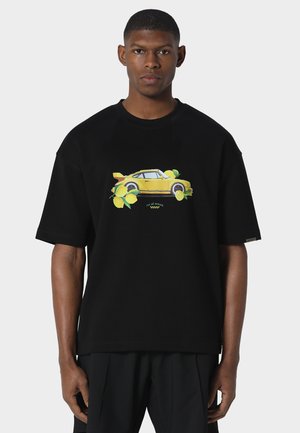 Jeune homme portant un T-shirt noir oversize avec une voiture de sport jaune et des citrons imprimés sur le devant, sur un fond uni.