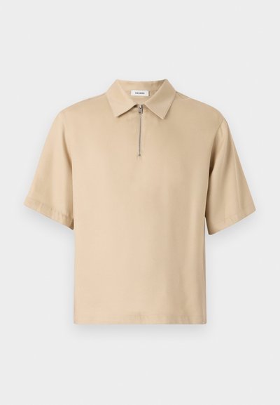 Μπεζ κοντομάνικο πόλο μπλουζάκι με γιακά και κοντό φερμουάρ στον λαιμό, φτιαγμένο από λεία ύφανση με χαλαρή εφαρμογή.