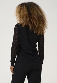 Top nero in pizzo a maniche lunghe con un motivo floreale testurizzato, abbinato a pantaloni neri. Il tessuto presenta un design semi-trasparente.