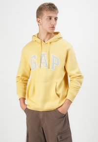 Geltona pralaidūs gobtuvas su pilku "GAP" logotipu, su priekinėmis kišenėmis, raišteliu ir guminiais rankogaliais. Dėvimi su rudais cargo šortais.
