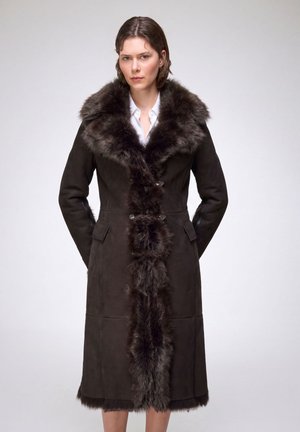 Femme portant un long manteau marron foncé avec une épaisse bordure en fourrure autour du col, sur le devant et à l'ourlet, debout devant un fond uni.