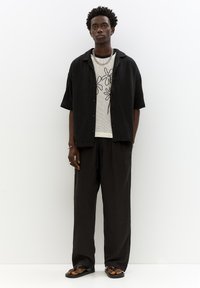 Camicia nera a maniche corte con bottoni sopra una maglietta bianca in rete con disegni floreali; abbinata a pantaloni neri larghi e infradito.