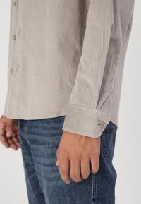Camisa de corduroy beige claro con rayas verticales, puños abotonados y tapeta de botones; combinada con pantalones vaqueros de mezclilla azul oscuro.