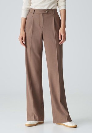 Woman wearing high-waist, wide-leg brown trousers with a front button, paired with white sneakers and a cream long-sleeve top.