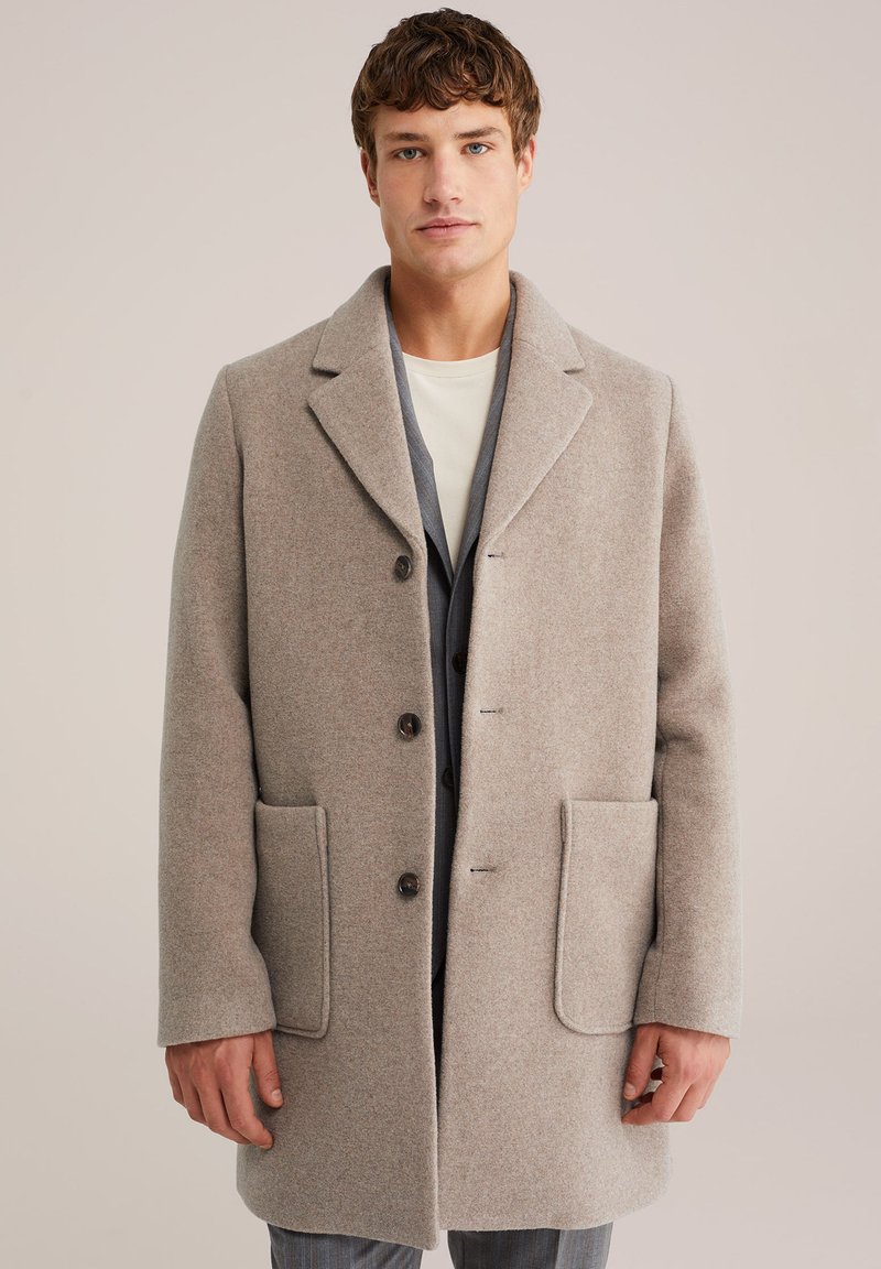 Jonge man in een beige wollen jas met grote zakken, knopen ervoor, en een grijze blazer eronder tegen een effen lichte achtergrond.