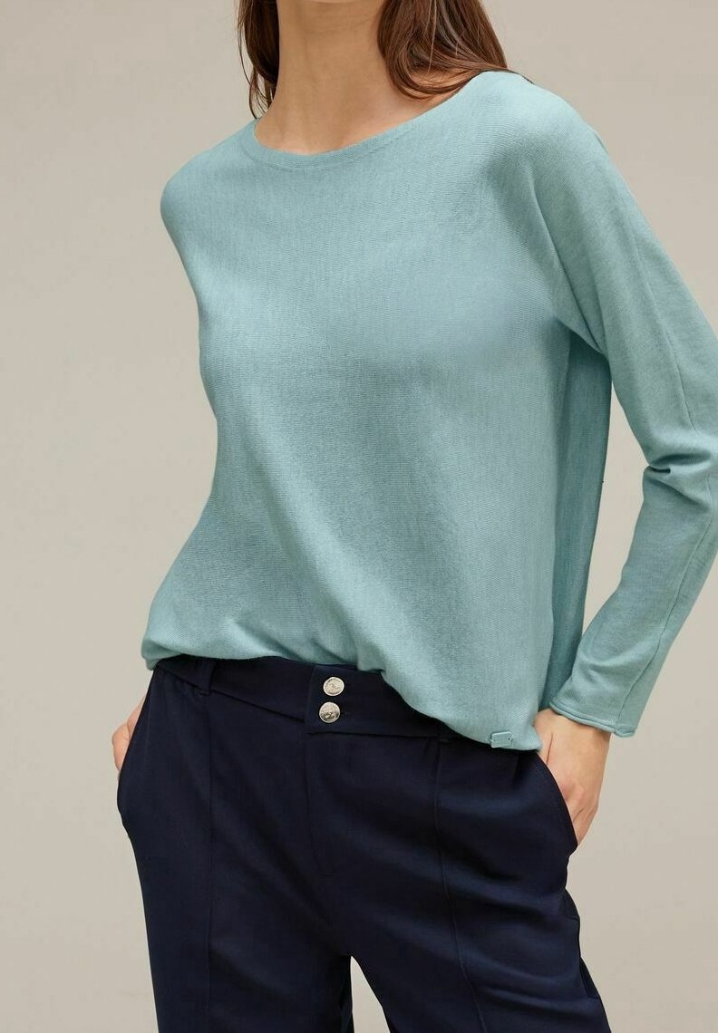 Femme portant un pull bleu clair à manches longues rentré dans un pantalon taille haute bleu marine avec deux boutons argentés, mains dans les poches.
