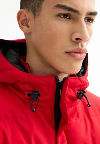 Joven vestido con una chaqueta con capucha roja, cordones ajustables negros y una cremallera etiquetada como "INSIDE" en el cuello.