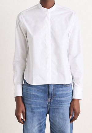 Person trägt ein weißes langärmliges Hemd mit Stehkragen, das in blaue, hoch taillierte Jeans gesteckt ist, vor einem einfarbigen Hintergrund.