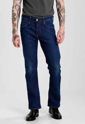 Man die donkere, rechte jeans draagt, zwarte laarzen en een donkergrijs T-shirt, staand tegen een effen lichte achtergrond.