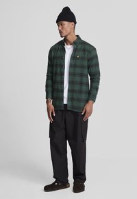 Camicia di flanella verde a scacchi con design a bottoni, indossata sopra una maglietta bianca. Pantaloni cargo neri e pantofole marroni completano l'outfit.