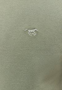 Lysegrøn tekstureret stof med et lille hvidt broderet hestelogo på brystet. Materialet ser blødt og strikket ud.