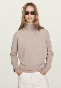 Vrouw in een beige coltrui, witte broek en zwarte rechthoekige zonnebril, staand met handen in de zakken tegen een effen achtergrond.