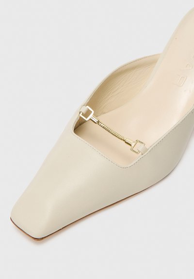Mule couleur crème avec un bout carré, texture en cuir lisse et un détail en chaîne dorée à l'ouverture. Doublure intérieure lisse.