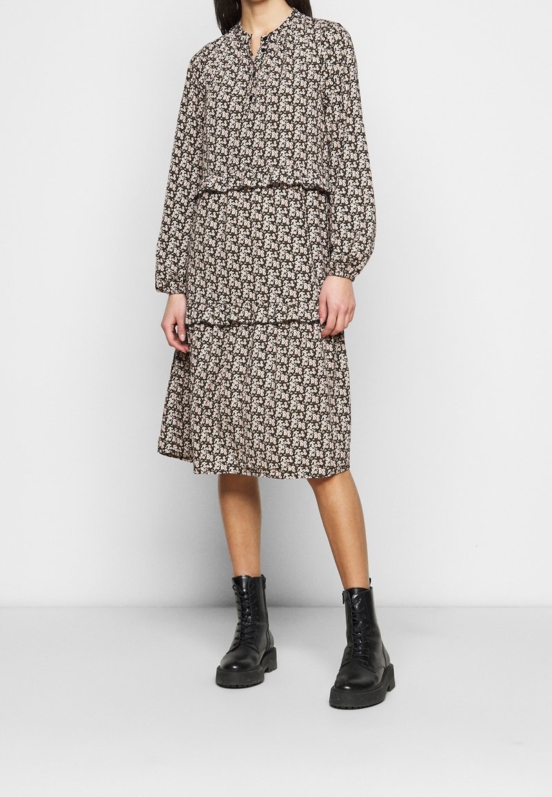 Květinové dlouhé šaty v tmavě hnědé barvě, s volánkovou sukní a nařaseným výstřihem; nošeny s černými bojovými botami.