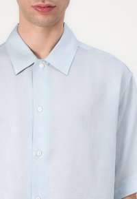 Camicia azzurro chiaro a maniche corte con un sottile motivo a foglia, tessuto texturizzato, chiusura con bottoni e colletto classico.