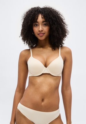 Γυναίκα με σγουρά μαλλιά που φοράει μπεζ σουτιέν και ταιριαστό εσώρουχο, κοιτώντας προς τα εμπρός ενάντια σε λευκό απλό φόντο.