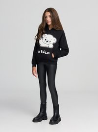 Felpa con cappuccio nera con grafica di un orso bianco e scritta "HELLO", indossata sopra leggings neri lucidi e stivaletti neri alla caviglia. Tessuto liscio, vestibilità comoda.