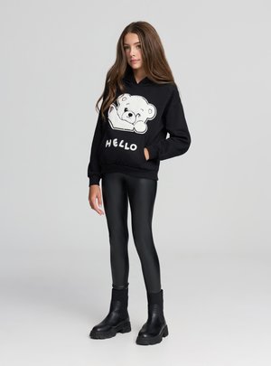 Felpa con cappuccio nera con grafica di un orso bianco e scritta "HELLO", indossata sopra leggings neri lucidi e stivaletti neri alla caviglia. Tessuto liscio, vestibilità comoda.
