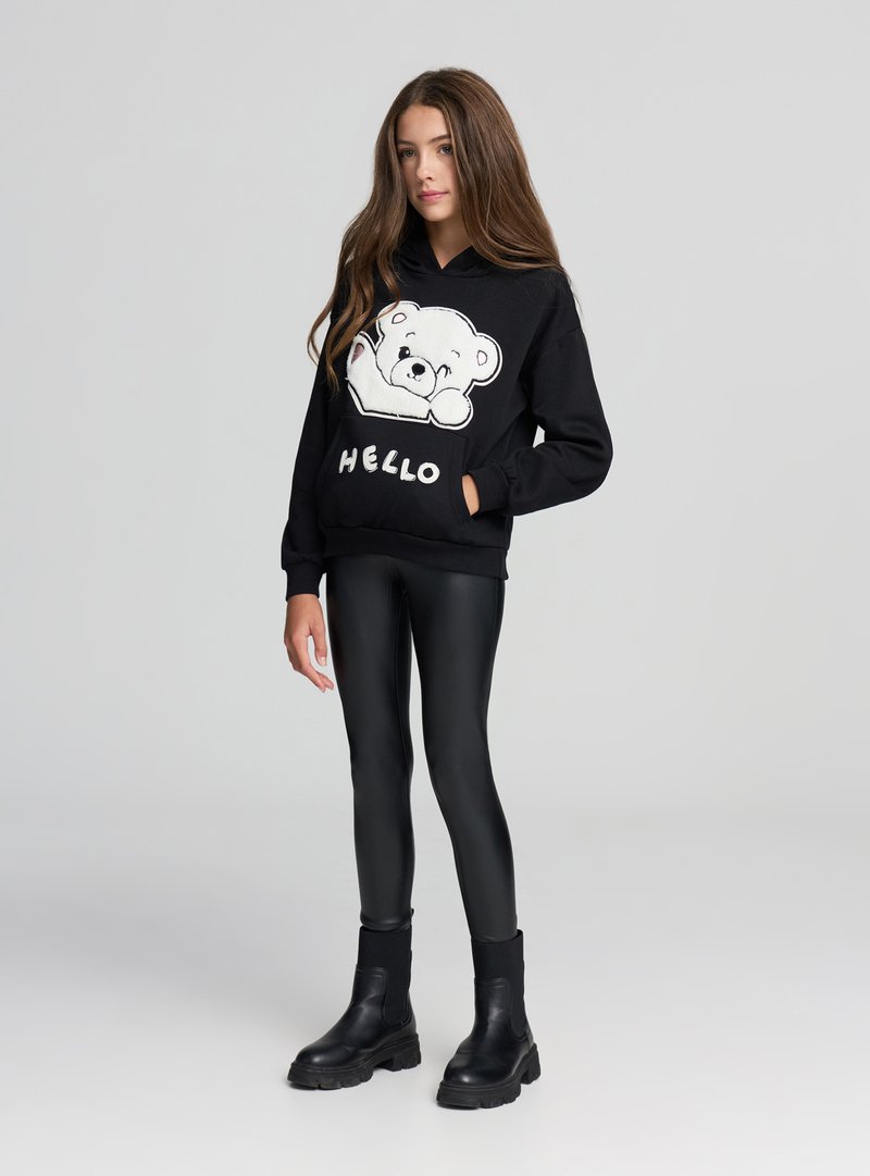 Felpa con cappuccio nera con grafica di un orso bianco e scritta "HELLO", indossata sopra leggings neri lucidi e stivaletti neri alla caviglia. Tessuto liscio, vestibilità comoda.