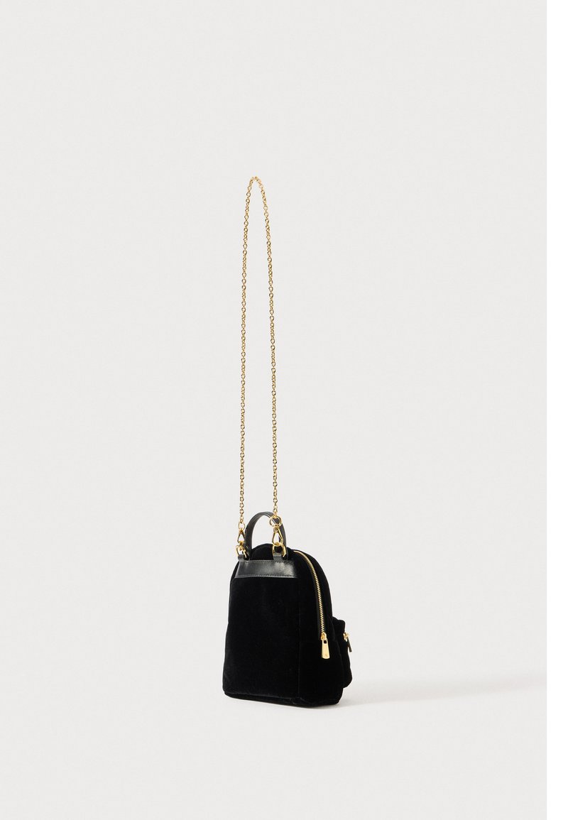 Maza melna samta plecu soma ar zelta rāvējslēdzēju un ķēdes siksnu, kā arī ādas rokturi uz viendabīga balta fona.