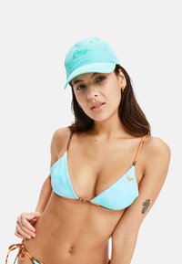 Lichtblauwe bikini met tropische bladerenpatronen, oranje accenten op de bandjes en een bijpassende turquoise pet met geborduurd logo.
