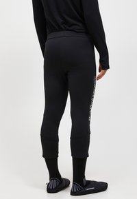 Schwarze, eng anliegende Sportleggings mit glatter Textur und einem weißen Seitenlogo. Kombiniert mit schwarzen Schuhen und gemusterten Socken.