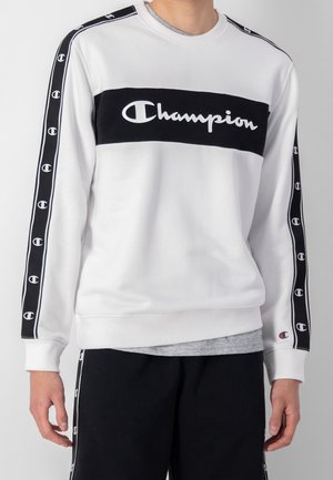 Osoba nosząca biały sweatshirt Champion z czarnym paskiem z logo na klatce piersiowej i czarnymi paskami z logotypami na rękawach.