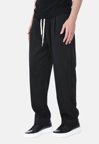 Pantaloni jogger neri realizzati in morbido tessuto, con vita elastica e cordino bianco, e tasche laterali. Indossati con sneakers nere.
