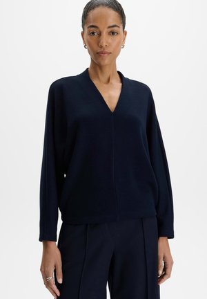 Woman wearing a dark navy long-sleeve V-neck top and matching pants stands against a plain white background.