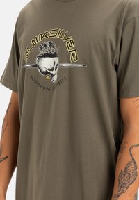 Camiseta verde oliva de algodón, con un gráfico de una calavera con palmeras y una tabla de surf. Diseño de manga corta y ajuste relajado.