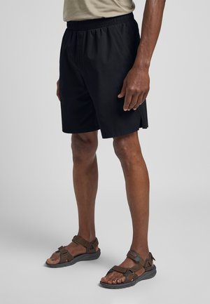 Schwarze Shorts mit elastischem Bund, seitlichen Schlitzen und einem glatten, leichten Stoff. Braune Sandalen mit verstellbaren Riemen und einer Gummisohle.