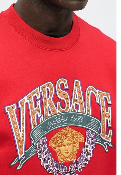 Punane kapuutsiga särk, millel on suur "VERSACE" logo kuldsete nikkudega, Medusa sümbol ja dekoratiivsed aktsendid lillades ja rohelistes toonides.
