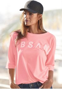 Light pink long-sleeve shirt with white text, a relaxed fit, and a round neckline. Paired with a black mesh baseball cap.