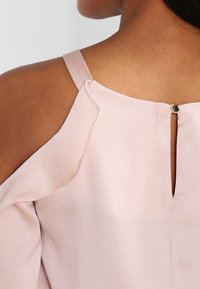 Blusa rosa chiaro con design a spalle scoperte, tessuto testurizzato e apertura a chiave sul retro con chiusura a bottone rotondo.