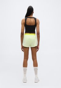 Γυναίκα όρθια με πλάτη προς τα εμπρός, φοράει μαύρο αμάνικο τοπ με άνοιγμα στην πλάτη, ανοιχτό κίτρινο σορτς, λευκές κάλτσες και λευκά αθλητικά παπούτσια σε απλό φόντο.