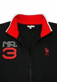 Svart zip-up jacka med en röd- och vitrandig krage. Har röda broderade "NR. 3" och "USPA" logotyper på framsidan. Mjuk tygstruktur.