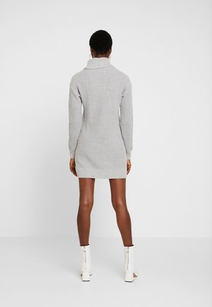 Femme portant une robe-pull en tricot gris clair et des bottines blanches, debout de dos sur un fond blanc uni.