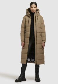 Beige pufferjas met een lange snit, zijzakken, een capuchon met trekkoord en geribbelde manchetten, gecombineerd met zwarte combatboots.
