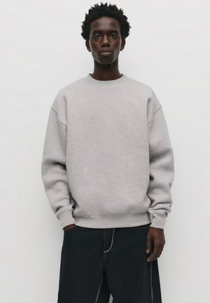 Szürke pulóver, kerek nyakkivágással és lejjebb ejtett vállakkal, puha anyagból készült. Sötét farmernadrággal párosítva, amelynek kontrasztos varrása van.