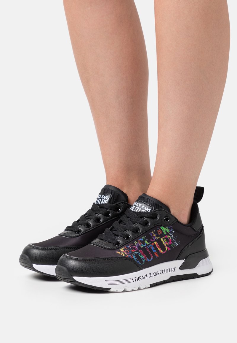 Mustat lenkkarit, joissa on synteettinen päällinen, valkoinen pohja ja monivärinen "VERSACE JEANS COUTURE" -teksti. Sisältää nauhat ja pehmustetun kauluksen.
