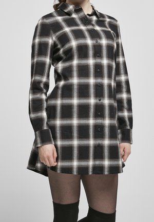 Vrouw in een zwart-witte geruite langwerpige blouse jurk met zwarte doorschijnende panty en zwarte kniekousen.