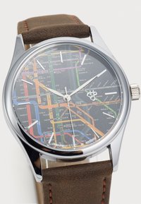 Watch with a round stainless steel case, black map dial featuring multi-colored subway lines, and brown leather strap with orange stitching.