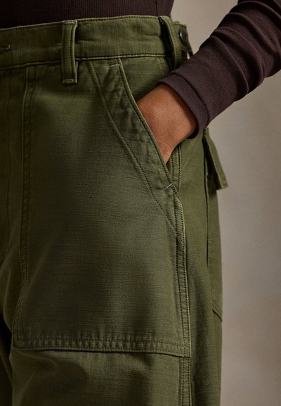 Παντελόνι cargo σε ελιά πράσινο με υφή υφάσματος, τονισμένες τσέπες και ορατές ραφές στη μέση και στις πλευρές.