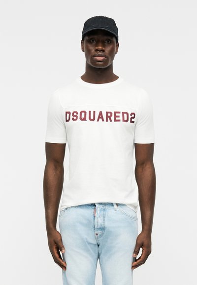 Άνδρας που φοράει λευκό t-shirt με το λογότυπο Dsquared2, ανοιχτό μπλε τζιν και μαύρο καπέλο μπέιζμπολ, στέκεται μπροστά σε ένα απλό φόντο.