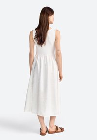 Vestido blanco con un torso ajustado y una falda fluida que llega hasta la mitad de la pantorrilla, complementado con sandalias marrones con un lazo decorativo.