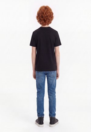 Rødhåret barn, der står med ryggen mod, iført en sort t-shirt, blå jeans og sorte høje sneakers på en hvid baggrund.