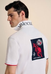 Camisa polo blanca con cuello pronunciado, marca azul, acentos rojos y un gran logo bordado que presenta a un jinete sobre un caballo.