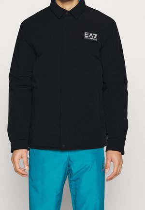 Mężczyzna ubrany w czarną kurtkę EA7 Emporio Armani i jaskrawo niebieskie spodnie, stojący z rękami swobodnie opuszczonymi wzdłuż ciała na jednolitym tle.
