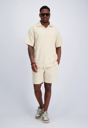 Man draagt beige polo met korte mouwen met textuur en bijpassende shorts, witte sneakers, zonnebril, staat met één hand in zijn zak.