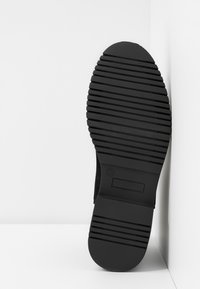 Semelle de chaussure noire avec des rainures horizontales appuyée contre un mur blanc d'angle et le sol, taille marquée 37 sur la semelle.